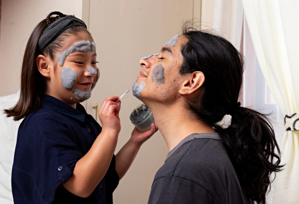 Young man with a girl sitting on a bed talking and laughing, while putting on a face cream, playing in a spa at home